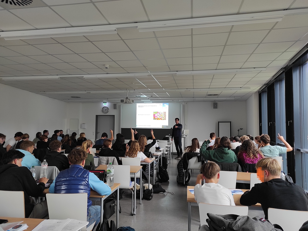 Hörsaal mit Schüler:innen von hinten mit Blick zur Tafel und zum Dozenten, einzelne melden sich