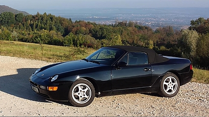 Porsche 968 in schwarz steht auf einem Berg, im Hintergrund ist ein Tal mit Bäumen und Wiese zu sehen.