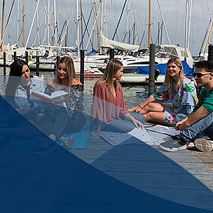 Studierende lernen und unterhalten sich auf einem Steg vor Booten bei sonnigem Wetter