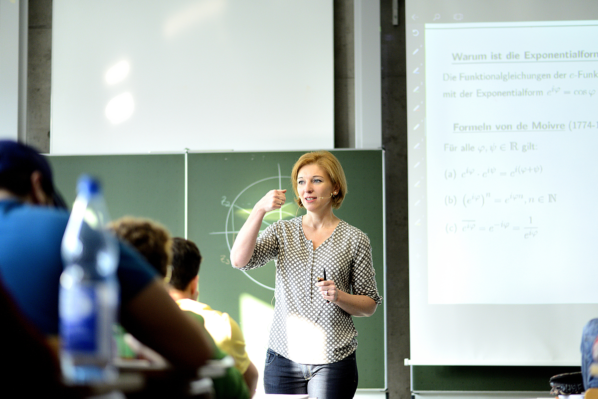 Weibliche Person steht in einem Hörsaal vor Studierenden und hält eine Vorlesung